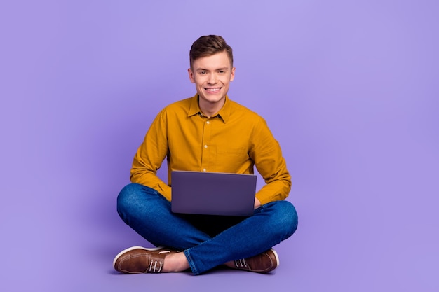 Full length photo of young cheeerful man use laptop marketer sit floor isolated over violet color background