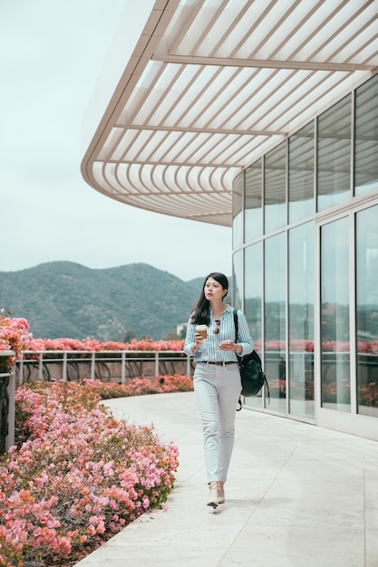 Full length portrait of attractive asian chinese businesswoman holding in hand paper cup of coffee and texting on mobile phone while walking in moring going to work. elegant lady on way to office.