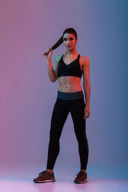 Full length portrait of attractive fit woman or sports trainer in sportswear posing  during fitness, isolated over purple wall