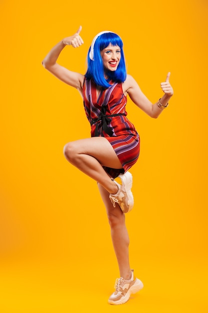 Full length portrait of an attractive funky young girl with blue hair wearing casual outfit jumping isolated over yellow wall, listening to music with headphones