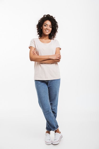 Full length portrait of beautiful american woman wearing jeans and t-shirt standing with arms folded and smile, isolated over white wall