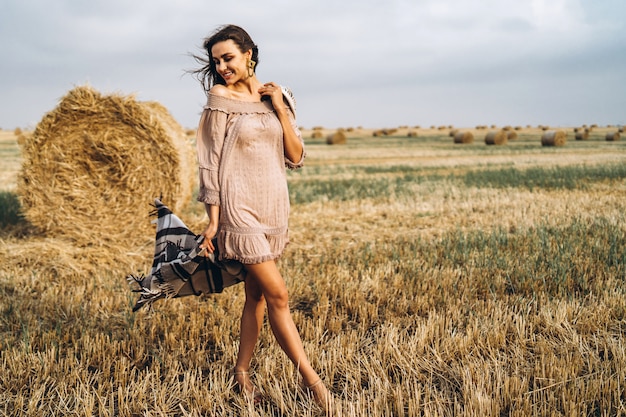 Full length portrait of a beautiful brunette in a dress and with a warm plaid. Woman enjoying a walk in a wheat field with hay bales