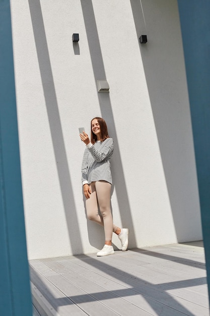 Full length portrait of beautiful winsome smiling woman wearing striped shirt using phone posing near house making selfie communication with friends online