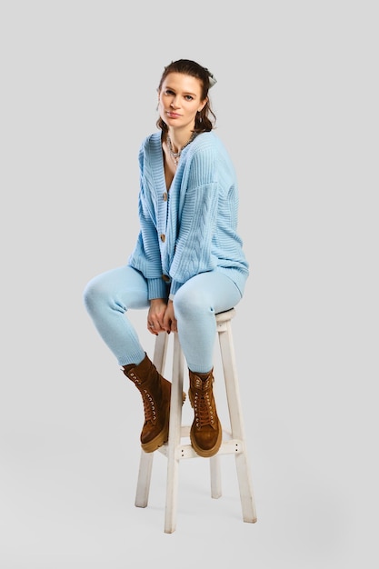 Full length portrait of beautiful woman in light blue warm knitted tracksuit sitting on stool over grey background