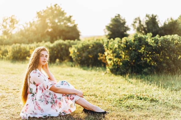 Full length portrait of beautiful woman with long curly hair on currant field background. Girl in a light dress sits on the grass in sunny day