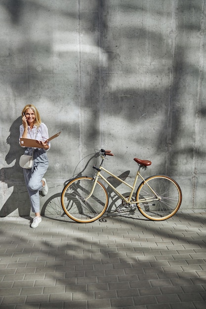 Full length portrait of charming blonde Caucasian woman holding paper folder with documents in hand while calling to her clients