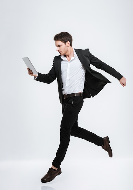 Full length portrait of a concentrated young business man jumping and looking at tablet computer isolated on a white wall