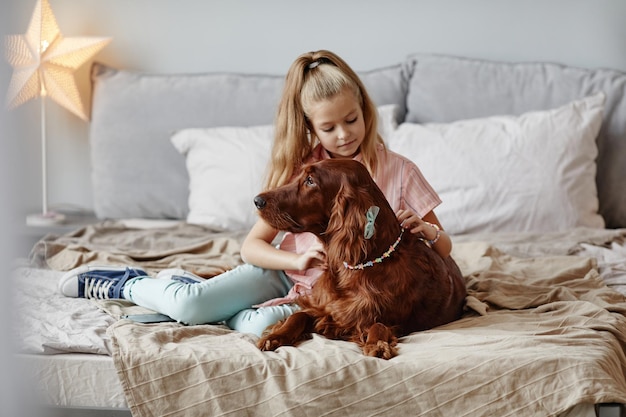 Full length portrait of cute blonde girl playing with dog on bed in cozy home interior, copy space