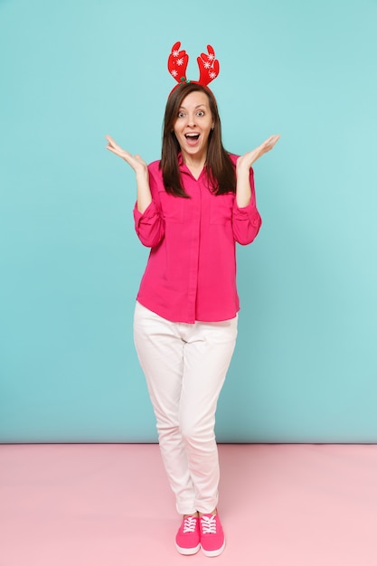 Full length portrait of fun young woman in rose shirt blouse, white pants, deer horns posing isolated on bright pink blue pastel wall.