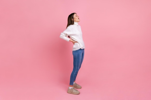 Full length portrait of girl touches injured back, suffers pain in lower lumbar muscular, arthritis, wearing white casual style sweater. Indoor studio shot isolated on pink background.