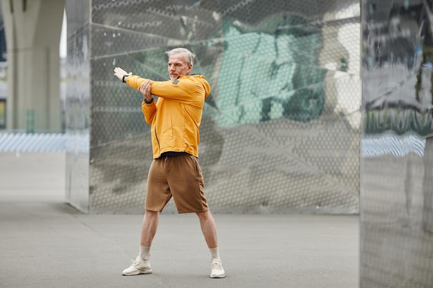 Full length portrait of handsome mature man doing stretching exercises outdoors in urban city settin