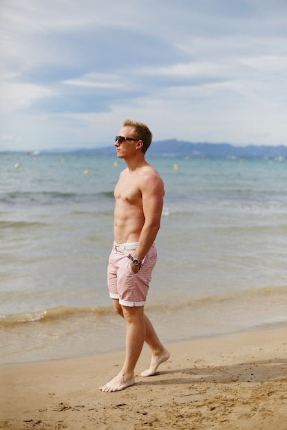 Full-length portrait of a handsome young man in beach shorts walking on the beach. Travel fashion. Sport lifestyle