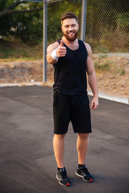 Full length portrait of a happy bearded fitness man showing thumb up outdoors