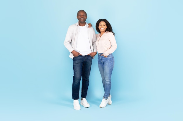 Full length portrait of happy black couple smiling at camera and posing standing over blue studio wall copy space