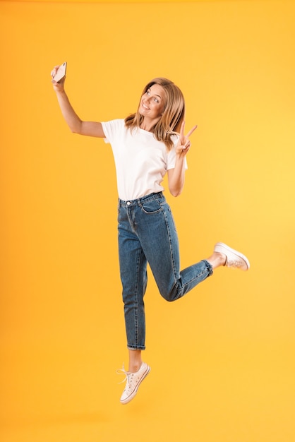 Full length Portrait of happy blond woman showing peace fingers while taking selfie Portrait isolated over yellow wall