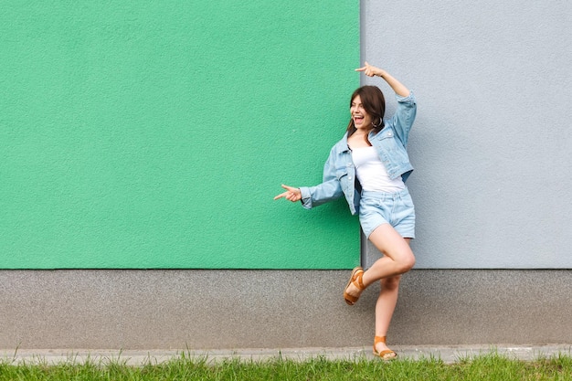 Full length portrait of happy excited beautiful woman in casual jeans denim style in summertime standing near green and light blue wall and pointing at green copyspace with two hands.