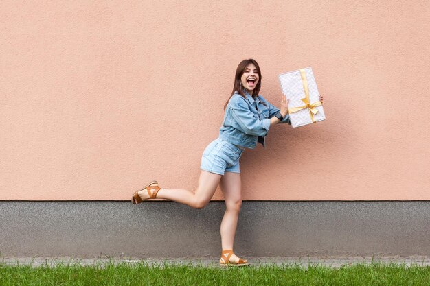 Full length portrait of happy excited beautiful woman in casual jeans denim style in summertime standing near sandybrown wall copyspace, holding gift box and looking at camera with surprised face.