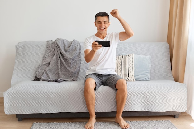 Full length portrait of happy excited smiling man wearing casual style white t shirt sitting on sofa completed level clenched fist celebrating victory expressing happiness