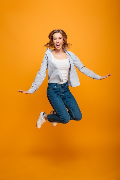 Full length portrait of happy joyous woman wearing braces shouting and having fun while jumping or levitating, isolated over yellow background