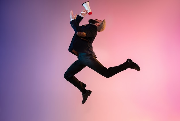 Full length portrait of happy jumping man wearing office clothes in neon light isolated on gradient background. Emotions, ad. Using mouthpeace, winning, sale, shopping, hurrying up, office work.