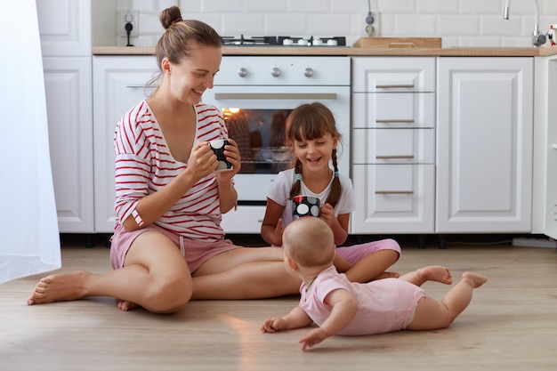 Full length portrait of happy mother wearing casual striped shirt sitting at floor and drinking hot coffee or tea while spending time with her daughters posing near woman.