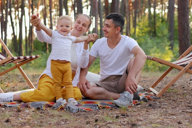 Full length portrait of happy positive woman and man sitting on ground in forest and playing with their toddler daughter couple spending weekend together enjoying beautiful nature and fresh air