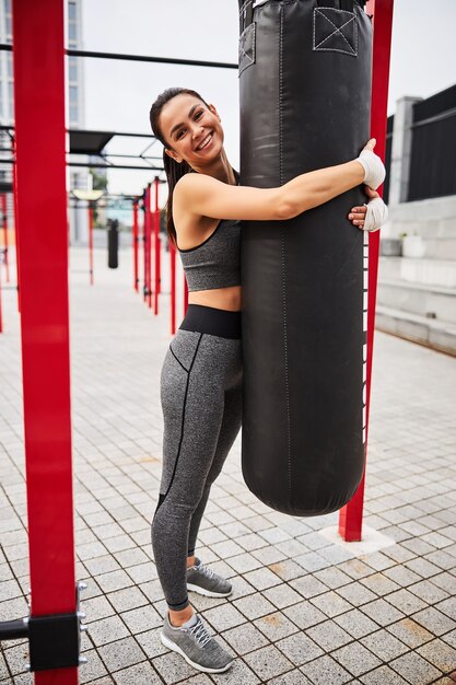 Full length portrait of joyful athletic female standing on playground and embracing outfit after workout