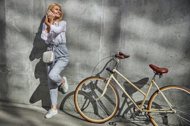 Full length portrait of laughing female standing near her bicycle while having conversation with business partner on mobile phone