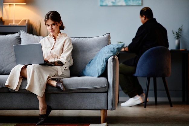 Full length portrait of modern young woman relaxing on couch at home and using laptop copy space