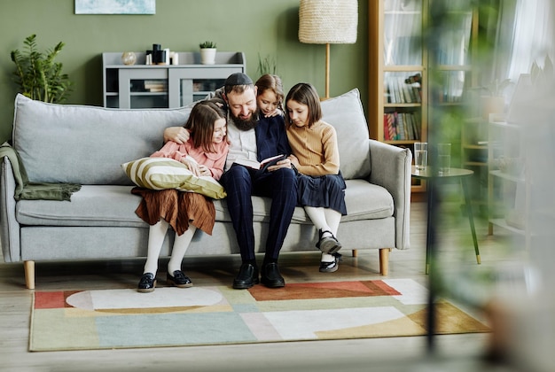 Full length portrait of orthodox jewish father reading book to three children at home copy space