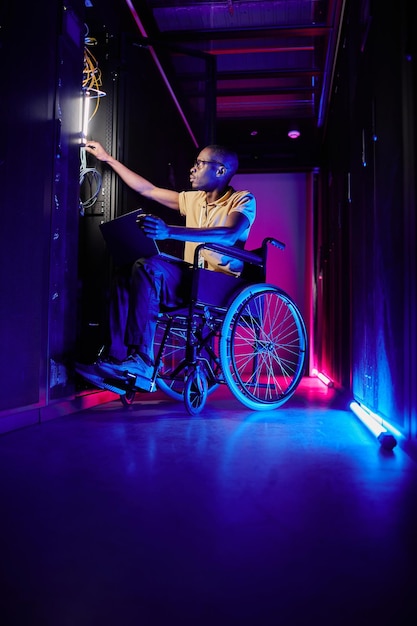 Full length portrait of person with disability working at system administrator in server room lit by