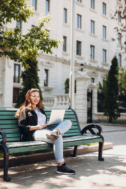 Full length portrait of a plus size woman sitting on a bench against a building with a laptop on her legs looking directly laughing.