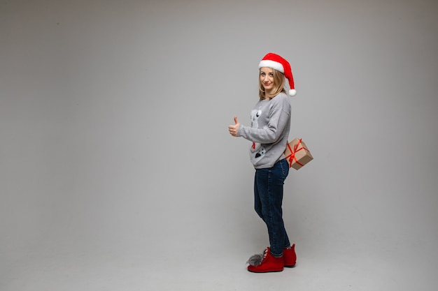 Full length portrait of pretty blonde woman in red Santa hat, sweater, jeans and boots holding gift behind her back and showing thumb up in approval