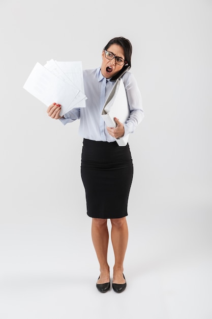 Full length portrait of a pretty busy businesswoman standing isolated over white wall, talking on mobile phone, holding folders