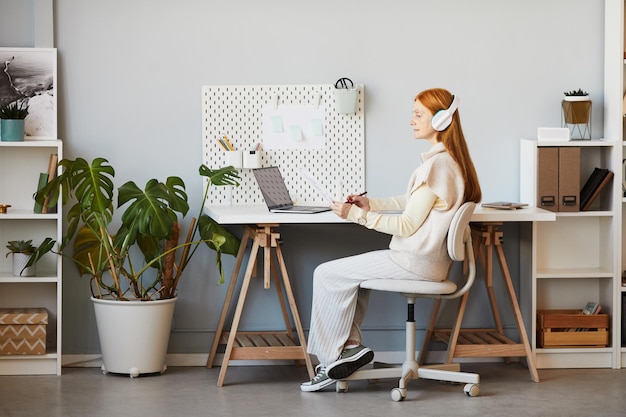 Full length portrait of red haired woman using laptop or studying at minimal home office workplace c...