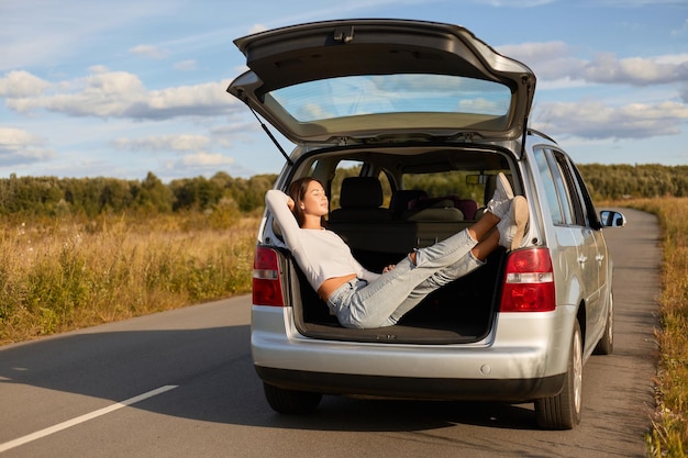 Full length portrait of relaxed woman sitting in the open trunk of a car and basking in the sun female wearing white shirt and jeans beautiful female traveling alone