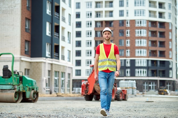 Full-length portrait of a serious calm young male worker in a protective helmet and a safety vest walking ahead