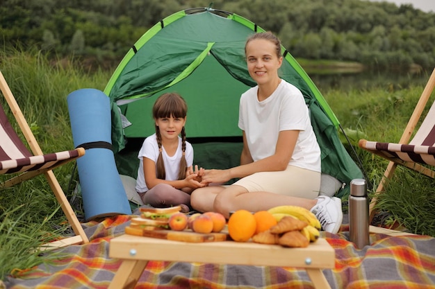 Full length portrait of smiling beautiful woman and little girl sitting on blanket near tent eating fruits and sandwiches having picnic enjoying warm summer day