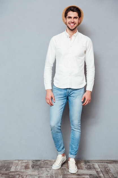 Full length portrait of smiling handsome young man in white shirt, jeans and hat standing