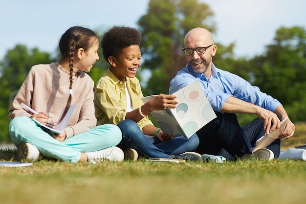 Full length portrait of smiling male teacher talking to children while sitting on green grass and enjoying outdoor class in sunlight