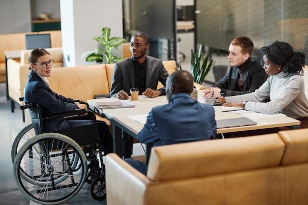 Full length portrait of successful businesswoman using wheelchair at meeting and talking to colleagues in modern office space