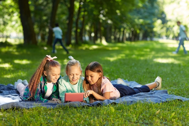 Full length Portrait of three teenage girls using digital tablet while lying on green grass in park outdoors lit by sunlight