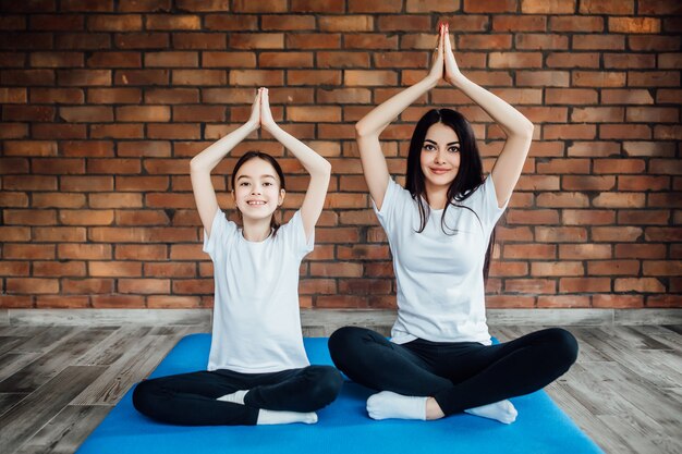 Full length portrait of two attractive girls  working out at home, doing yoga exercise on blue mat, sitting in Easy, on blue mat