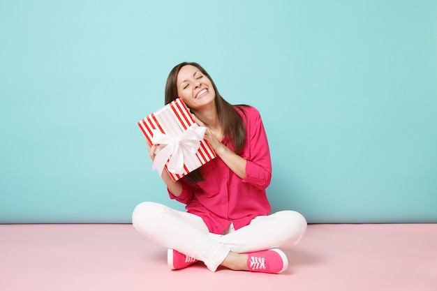 Full length portrait woman in rose shirt blouse white pants sitting on floor hold present box 