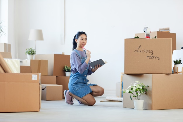 Full length portrait of young Asian woman organizing moving in process while sitting on floor next to cardboard boxes and holding planner