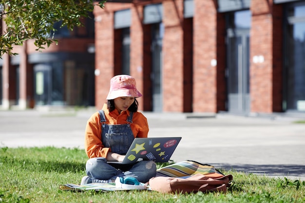 Full length portrait of young Asian woman using laptop sitting on grass outdoors in college campus