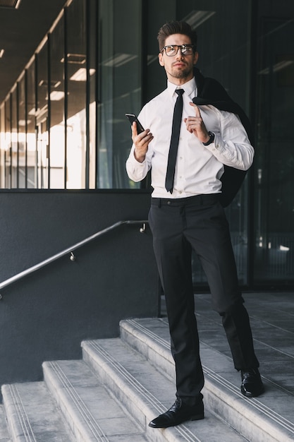 Full length portrait of young entrepreneur dressed in formal suit standing outside glass building with jacket over his shoulder, and holding mobile phone