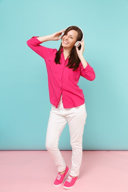 Full length portrait of young fun woman in rose shirt blouse, white pants, headphones dancing isolated on bright pink blue pastel wall..