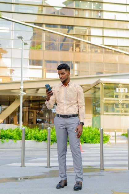 Full length portrait of young handsome African businessman in city using phone vertical shot