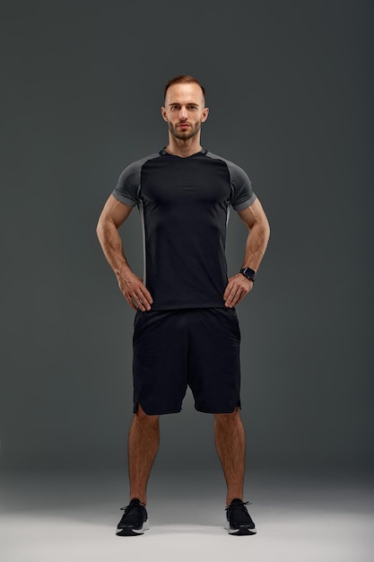 Full length portrait of young sporty guy in tshirt and shorts standing on gray studio background Serious millennial sportsman looking at camera Healthy lifestyle and sports concept